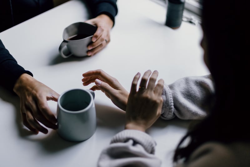 A hand pushing a coffee cup towards someone across a table, while holding a cup in the other hand